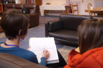 Caseworker sits on sofa with a woman. They are going through paperwork. Their backs are to the camera